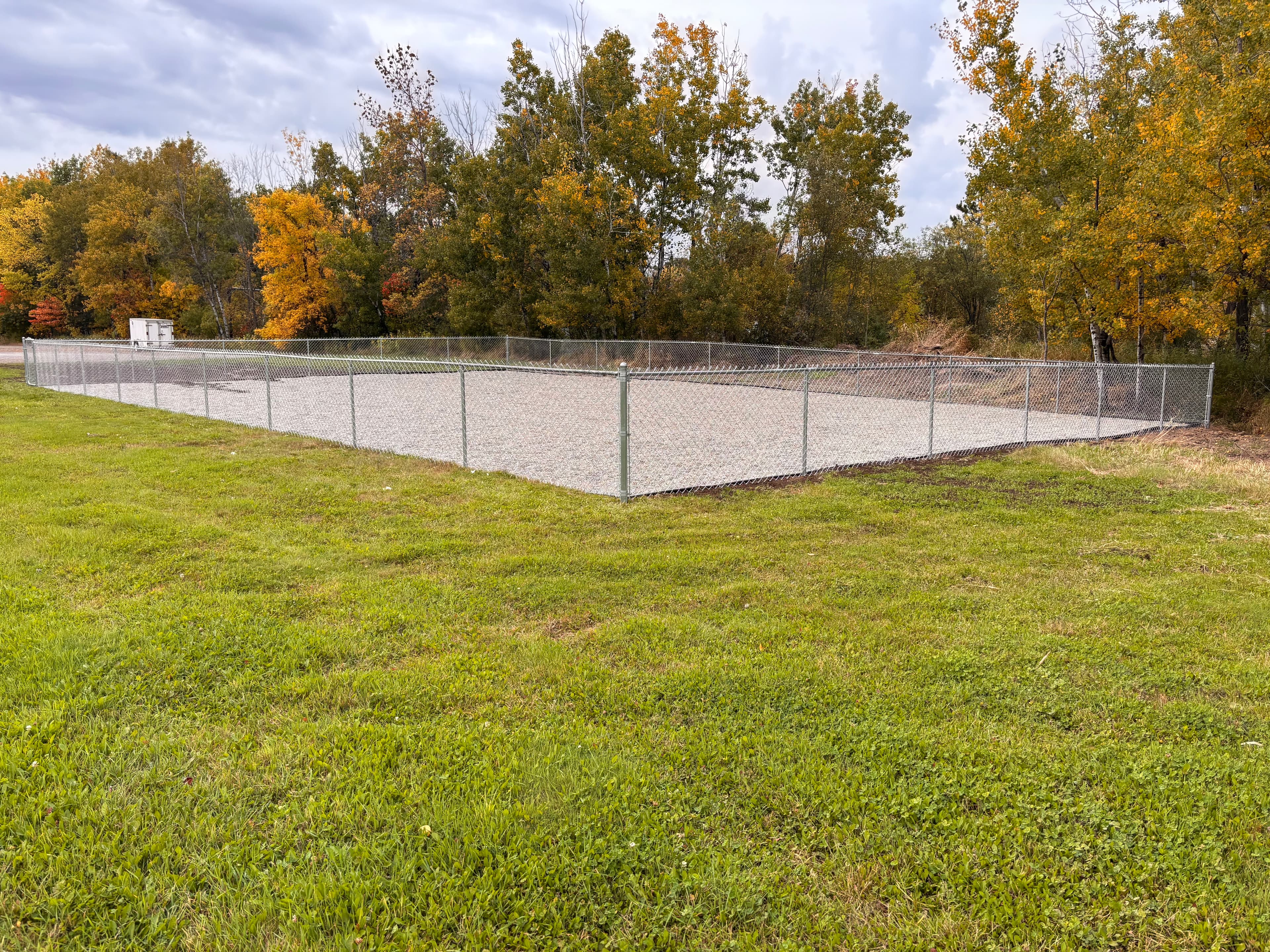 Chain-link fence enclosing a gravel lot in a grassy field with colorful autumn trees.