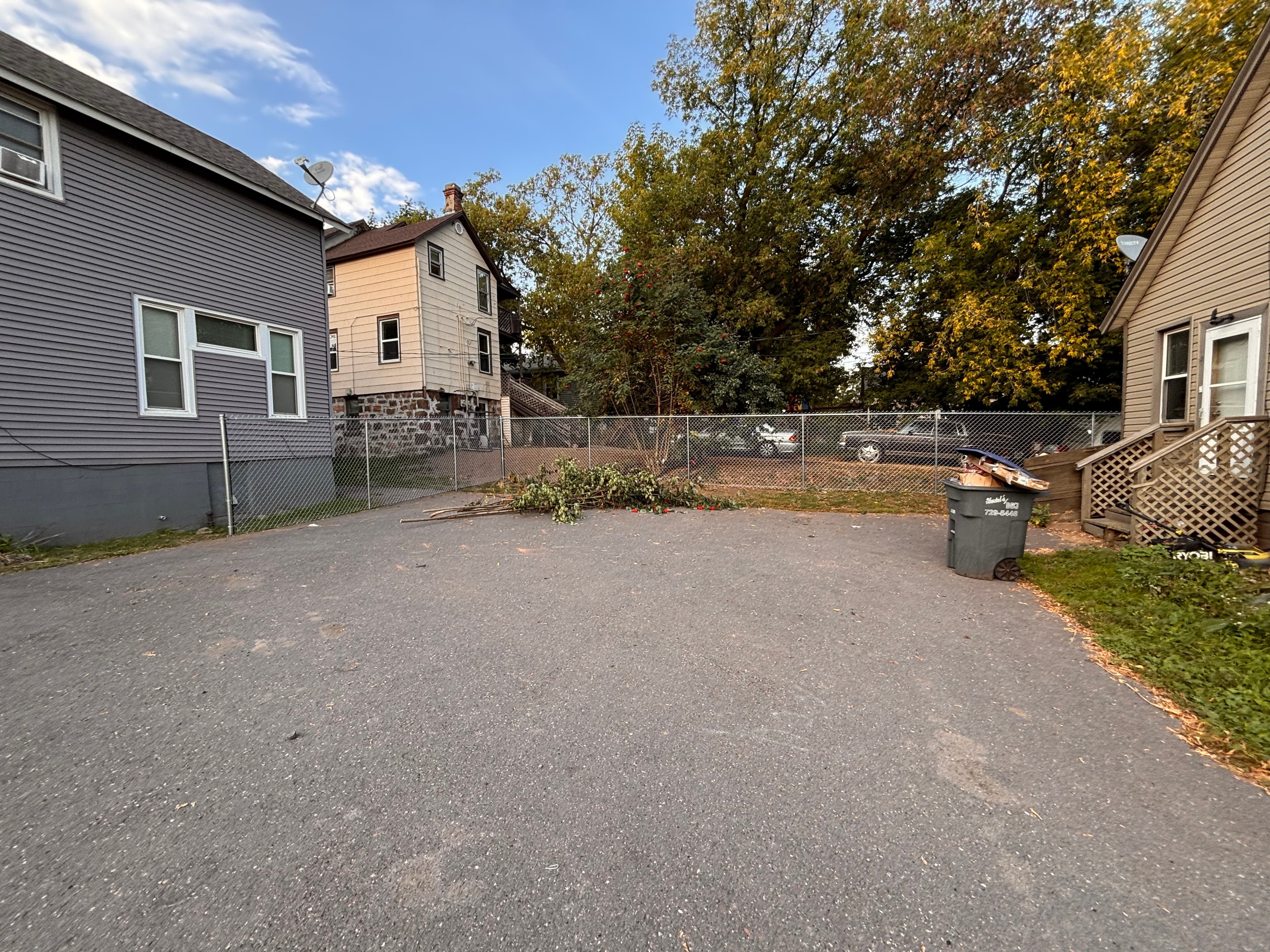 Asphalt driveway between two houses with cut tree branches and an overflowing trash bin.