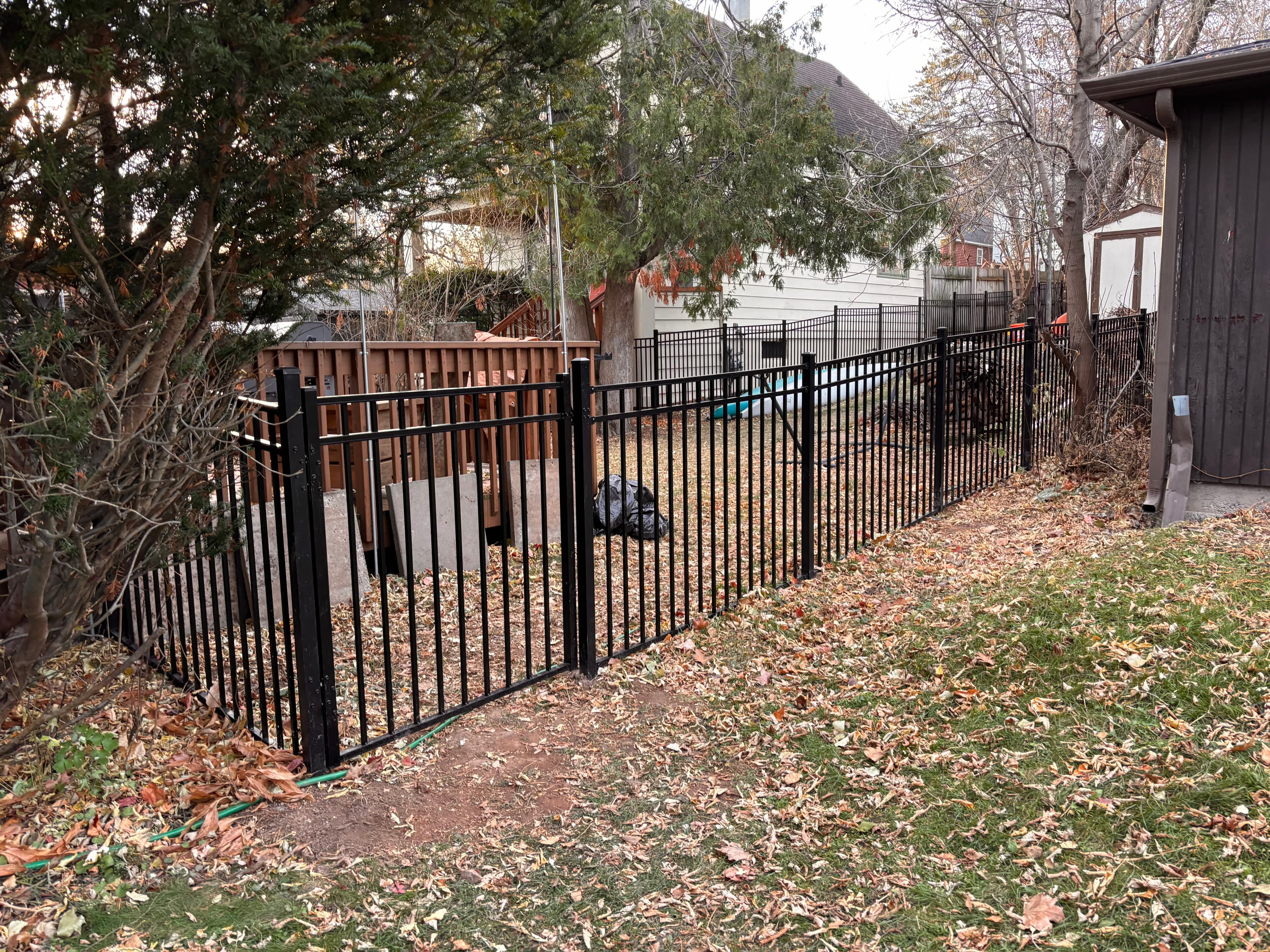 Black metal fence in a backyard covered with autumn leaves, featuring a wooden deck.